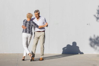 Sokakta beyaz bir duvarın önünde tango yapan mutlu emekli çift.