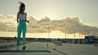 Gün batımı egzersizleri sırasında dinamik zıplama hareketleri yapan bir kadın. Sportif taytlar giyip şehir çatılarının tepesinde dramatik şehir manzarası var.