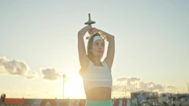 Atletik kadın, gün batımında çatıdaki bir antreman sırasında dambılla trisepsi uzatır.