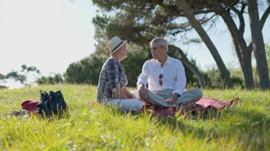 Yaşlı çift boş vakitlerin tadını çıkarıyor, doğanın ortasında piknik battaniyesinde sohbet ediyorlar.