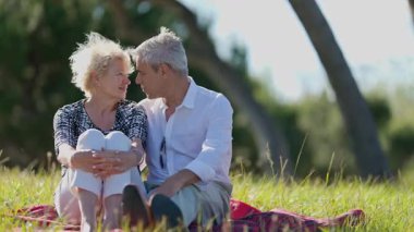 Yaşlı çift parkta battaniyede oturuyor, gülümsüyor ve birbirlerine eşlik ediyorlar.