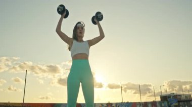 Atletik kadın gün batımında dışarıda halter egzersizleri yapıyor.