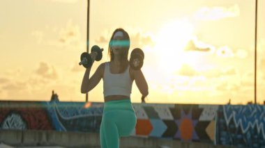 Genç bir kadın, günbatımının canlı arkaplanına karşı halterlerle güç alıştırması yapıyor.