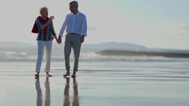 Mutlu yaşlı çift gün batımında sahilde yalınayak yürüyor.