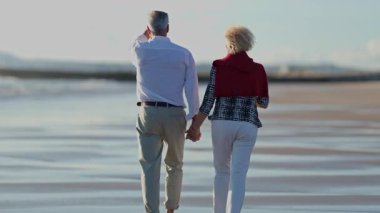 Yaşlı çift kumsalda rahatça yürüyüp el ele tutuşuyor ve emekliliklerinin tadını çıkarıyorlar.