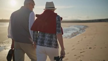 Gün batımında sahilde çıplak ayakla yürüyen yaşlı bir çift.