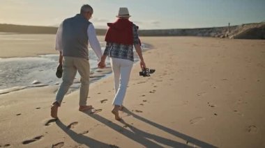 Gün batımında sahilde çıplak ayakla yürüyen yaşlı bir çift.