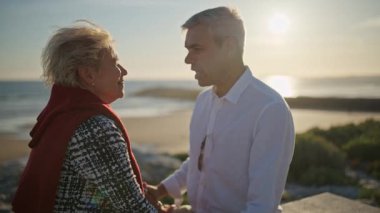 Romantik olgun çift gün batımında el ele tutuşup deniz kenarında konuşuyor.