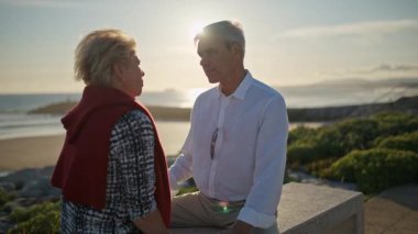 Yaşlı çift gün batımında okyanus kenarında bankta konuşmaktan zevk alıyor.