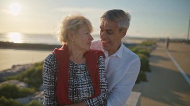 Deniz kenarında romantik bir anın tadını çıkaran yaşlı bir çift güzel bir gün batımında birbirlerine sarılıp gülümsüyorlar.