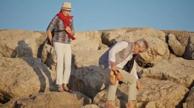 Yaşlı turistler sahilde kayalara tırmanırken birbirlerine yardım ediyorlar.