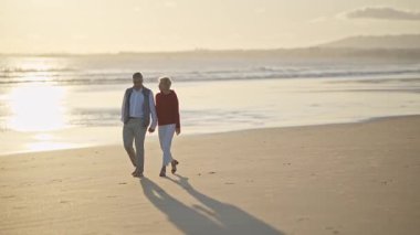 Yaşlı çift gün batımında sahilde romantik bir yürüyüş yapıyor.