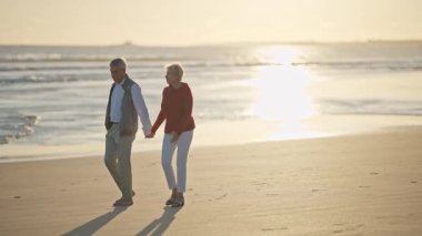 Yaşlı çift gün batımında sahilde romantik bir yürüyüş yapıyor.