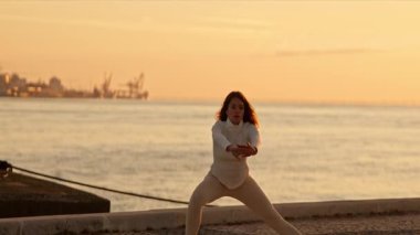 Kadın gün doğumunda denizin yakınındaki beton bir yüzey üzerinde nazikçe esneme egzersizleri yapıyor.