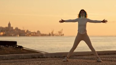 Kadın gün doğumunda nehir kenarında tai chi chuan yapıyor, arka planda bir şehir manzarası var.