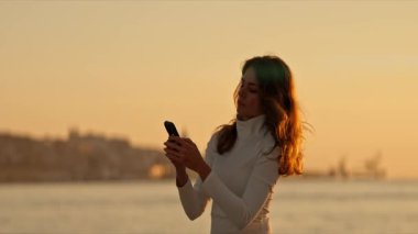 Kadın, gün batımında arka planda sakin bir şehir manzarası olan pitoresk bir rıhtımın yanındaki akıllı telefonunu kontrol ediyor.