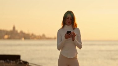 Günbatımında akıllı telefon kullanan kadın ve arka planda şehir manzarası