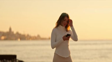 Şık bir kadın şehrin arka planında telefonunu kullanarak deniz kenarında altın saat geçirmekten hoşlanır.