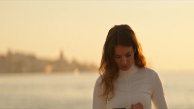Kulaklık takan ve güneş doğarken deniz kenarında akıllı bir telefon tutan genç bir kadın.