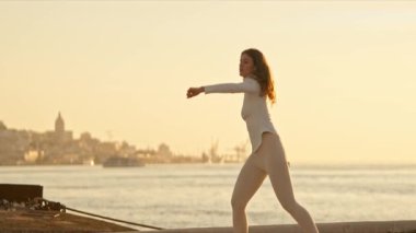 Şehir manzaralı bir iskelede modern bir bale gösterisi sergileyen zarif bir dansçı ve arka planda denizin üzerinde altın gün batımı.