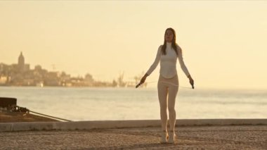 Spor salonundaki kadın gün batımında denizin önünde ip atlama antrenmanı yapıyor.