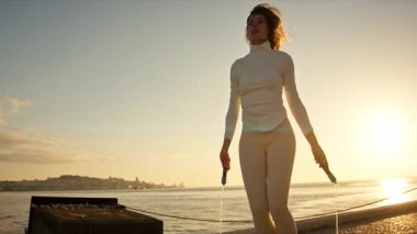 Zinde bir kadın güneş doğarken deniz kenarında atlama ipiyle egzersiz yapıyor.