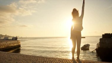 Gün batımında nehrin kenarında dans eden enerjik kadın, altın saatin tadını çıkarıyor.