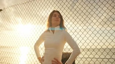 Gün batımında tel örgülerin yanında elleri kalçasında duran genç atletik bir kadın.