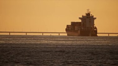 Altın bir günbatımında sakin sularda yol alan konteyner gemisi, arka planda görünen bir köprü ile