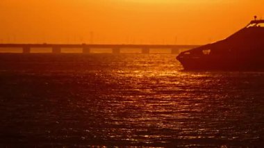 Suda portakal rengi gün batımı ve köprünün yanında giden yolcu feribotu silueti.
