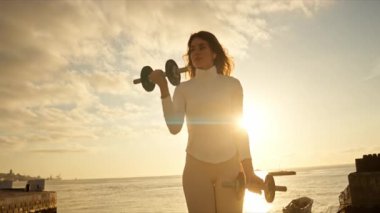 Fitness kadını gün doğumunda iskelede dambıllarla egzersiz yapıyor.