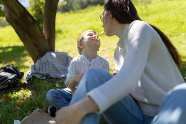 Anne ve oğul birlikte güzel vakit geçiriyorlar, güneşli bir piknik sırasında patates kızartması paylaşıyorlar, aile bağlarını kuvvetlendiriyorlar.