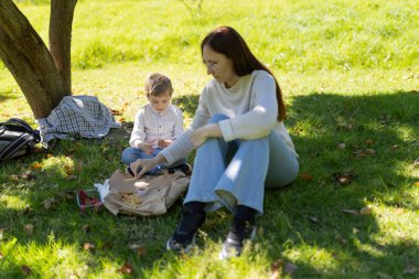 Anne ve oğul, güneşli bir sonbahar gününde yeşil bir parktaki ağacın gölgesinde yemek yiyorlar.