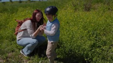 Kızıl saçlı anne çömelmiş, çayırda yeşil miğfer takan oğluna çiçekler gösteriyor.