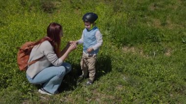 Anne ve oğul doğada bir anı paylaşıyor, güneşli bir çayırda çiçek topluyor.