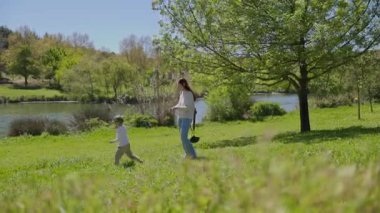 Mutlu bir aile parkta güneşli bir günün tadını çıkarıyor, gölün kenarında yürüyor ve ördekleri gözlemliyor.