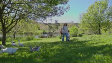 Anne ve oğlu güneşli bir günde göl kenarındaki kazların yanında parkta yürüyüş yapıyorlar.