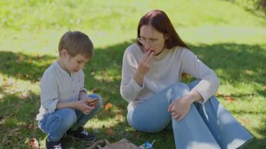 Anne ve oğul, güneşli bir günde huzur dolu bir park ortamında yiyecekleri paylaşırlar.