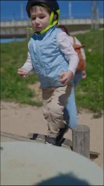 Miğferli bir çocuk kumlu bir yüzeyle karşılaşır, kayaları ve çiçekleri düzenler, açık havada merak ve konsantrasyon gösterir.