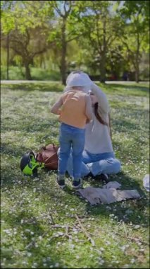 Anne oğluna parkta beyaz gömlek giydiriyor.