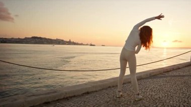 Kızıl saçlı kadın yoga yapıyor. Gün doğumunda Lizbon 'da bir rıhtımda poz veriyor.