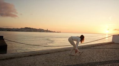 Zarif kadın güzel bir gündoğumu sırasında asfalt bir nehir kıyısında jimnastik hareketleri yapıyor.