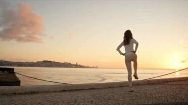 Gün doğumunda nehir kenarında ısınan genç bir kadın bacak esneme hareketleri yapıyor.