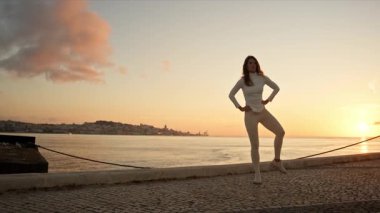 Dansçı, gün doğumunda nehir kenarında zarif pozlar vererek, manzaralı bir şehir zeminine karşı denge ve zarafet sergiliyor.