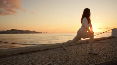 Lizbon 'daki Tagus nehrinin kıyısındaki kaldırımda gün doğumunda spor yapan kadın bacak esnetiyor.