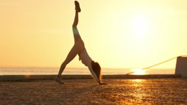 Kadın, gün doğumunda deniz kenarında asfalt bir yüzey üzerinde köpek yogasına bakan üç ayaklı bir sahne sergiliyor.