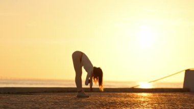 Sporcu kadın gün doğumunda sahilde eğilip egzersiz yapıyor.