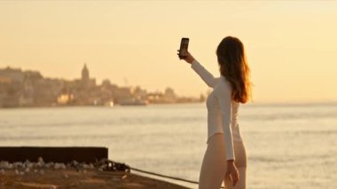 Genç bir kadın gün batımında Tagus nehri ile Lizbon 'da akıllı telefonuyla selfie çekiyor.