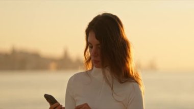 Gün batımında arka planda şehir manzarası olan cep telefonu kullanan genç bir kadın.