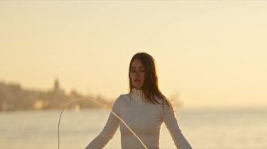 Gün batımında sakin bir rıhtımın yanında ustalıkla ip atlayan bir kadın. Manzaralı bir zemine karşı vücut bulmuş zindelik ve farkındalığı somutlaştırıyor.